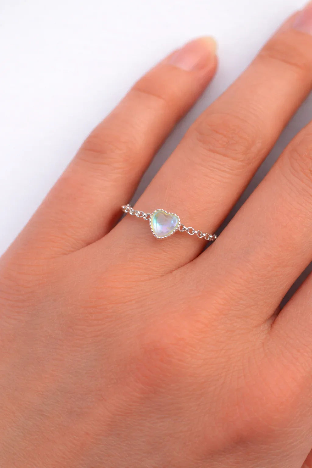 Main gauche avec bague fine en argent et pierre cœur irisée sur fond blanc